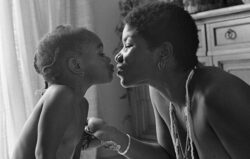 Black and white image of a Black mother with short hair leaning in to kiss her daughter in their living room.