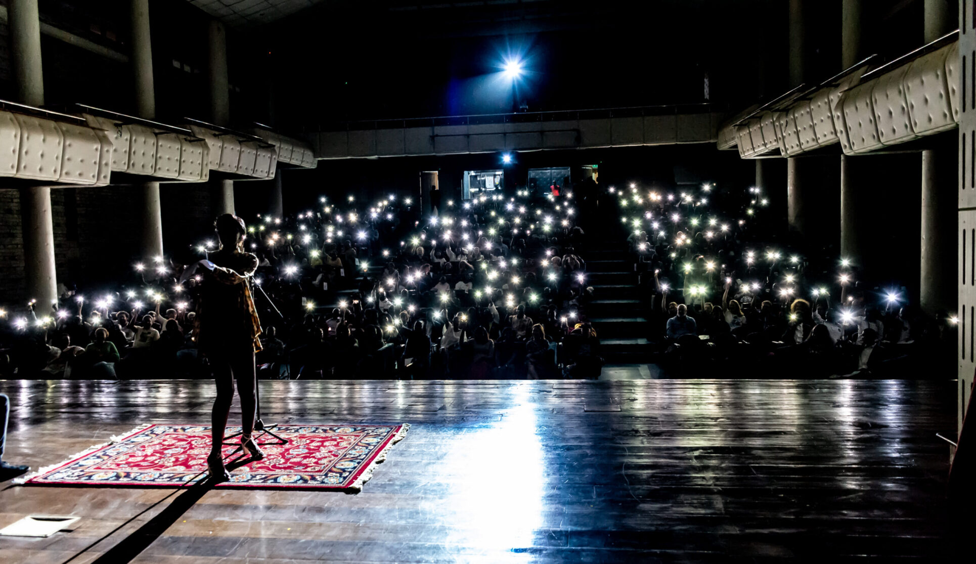 View from a stage our to a darkened audiorium with audience members holding up lights in the background.