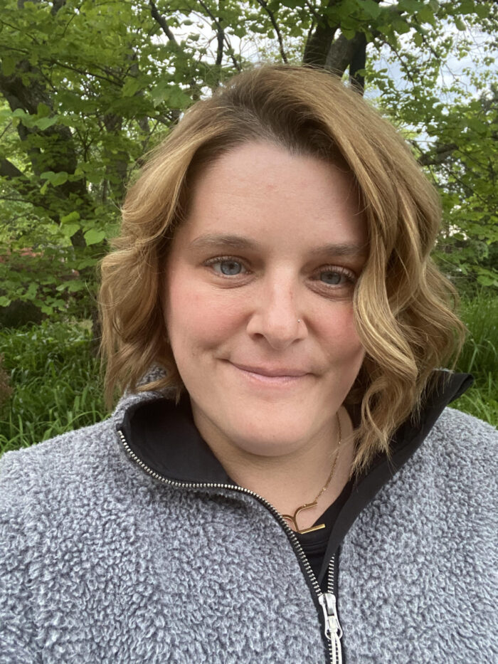 Ryann Morales wearing a grey sweater smiling with green leafed trees behind her.