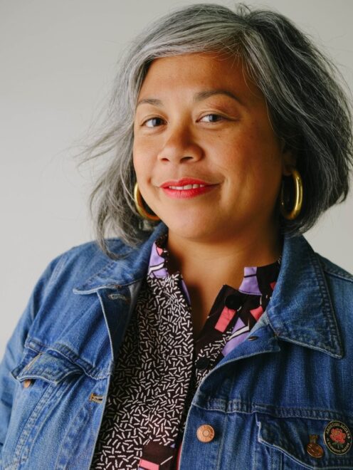 headshot of Angela wearing a denim jacket and smiling at the camera with a grey wall behind her.