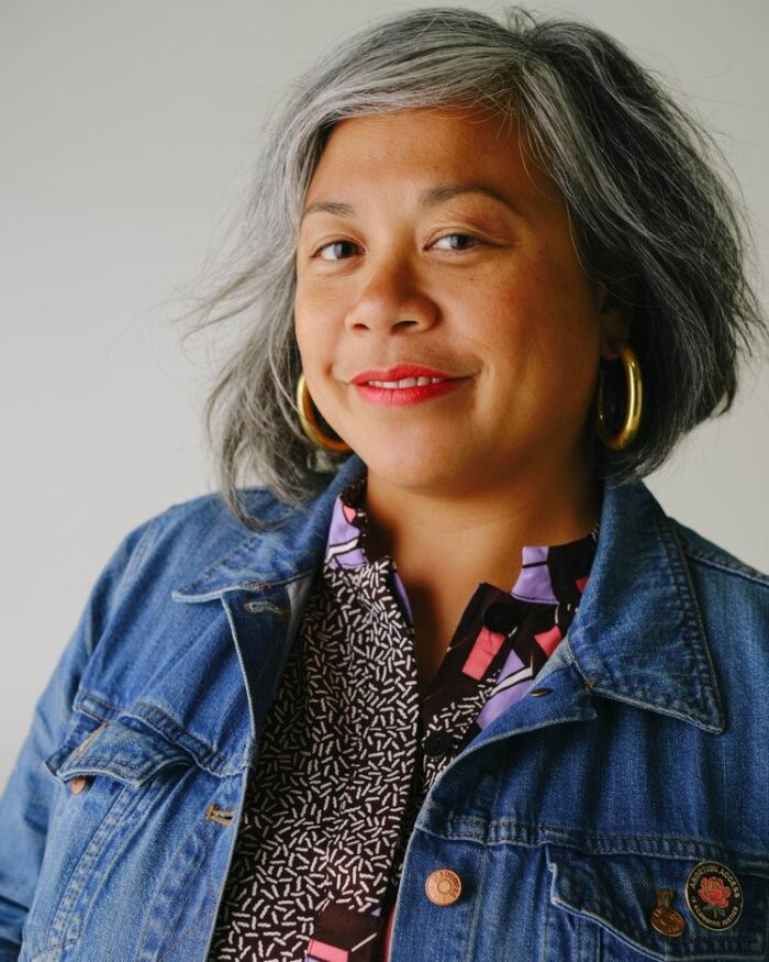 headshot of Angela wearing a denim jacket and smiling at the camera with a grey wall behind her.