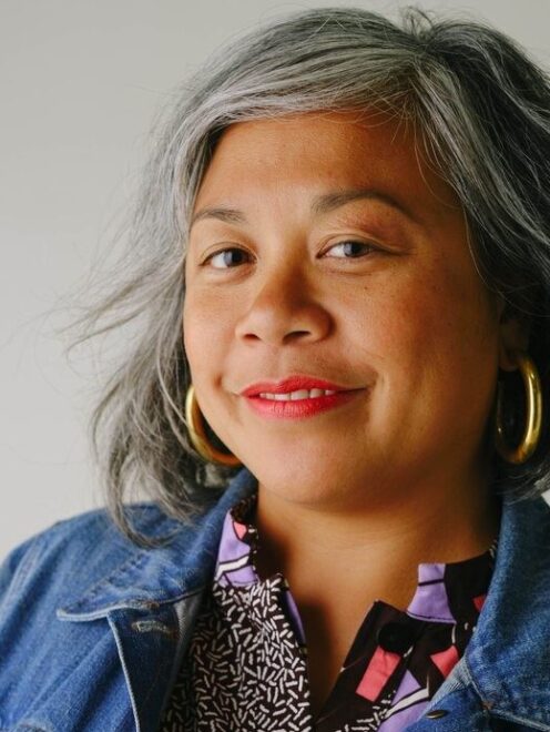 headshot of Angela wearing a denim jacket and smiling at the camera with a grey wall behind her.