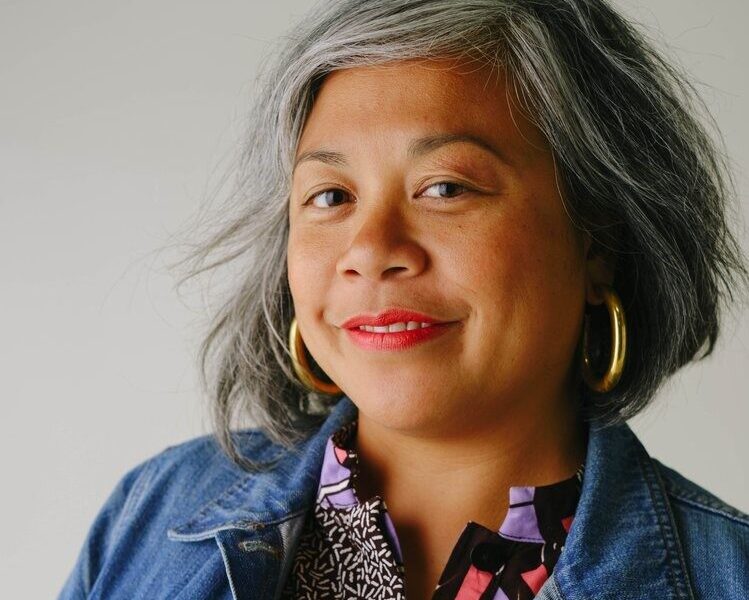 headshot of Angela wearing a denim jacket and smiling at the camera with a grey wall behind her.