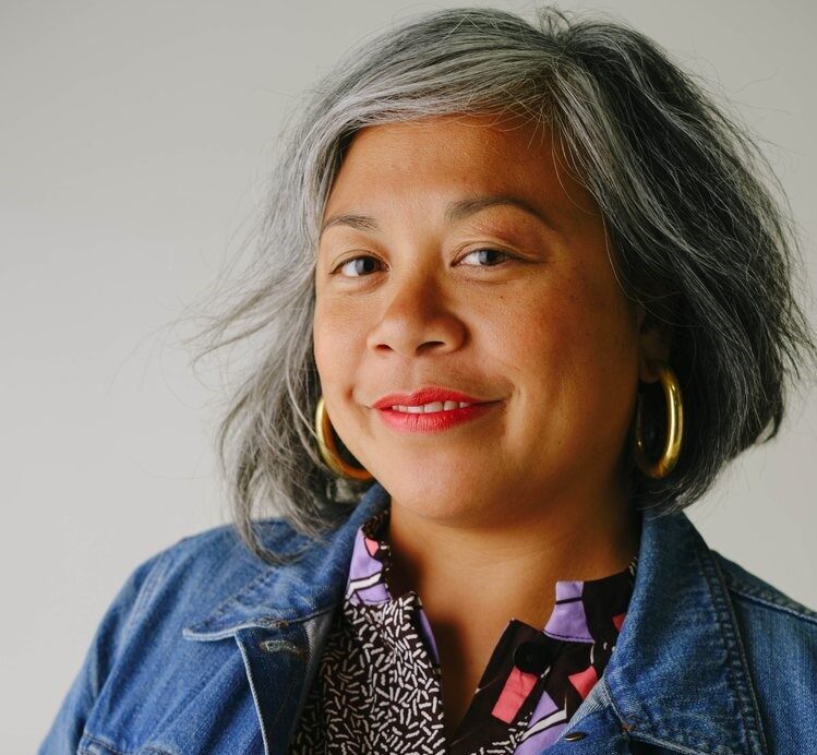 headshot of Angela wearing a denim jacket and smiling at the camera with a grey wall behind her.