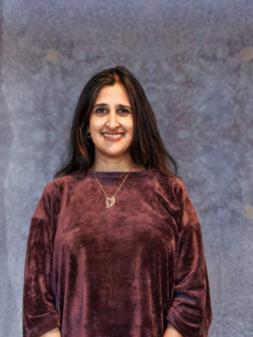 Dila stands in front of a grey backfround. She has dark hair and is wearing a maroon shirt and smiling.