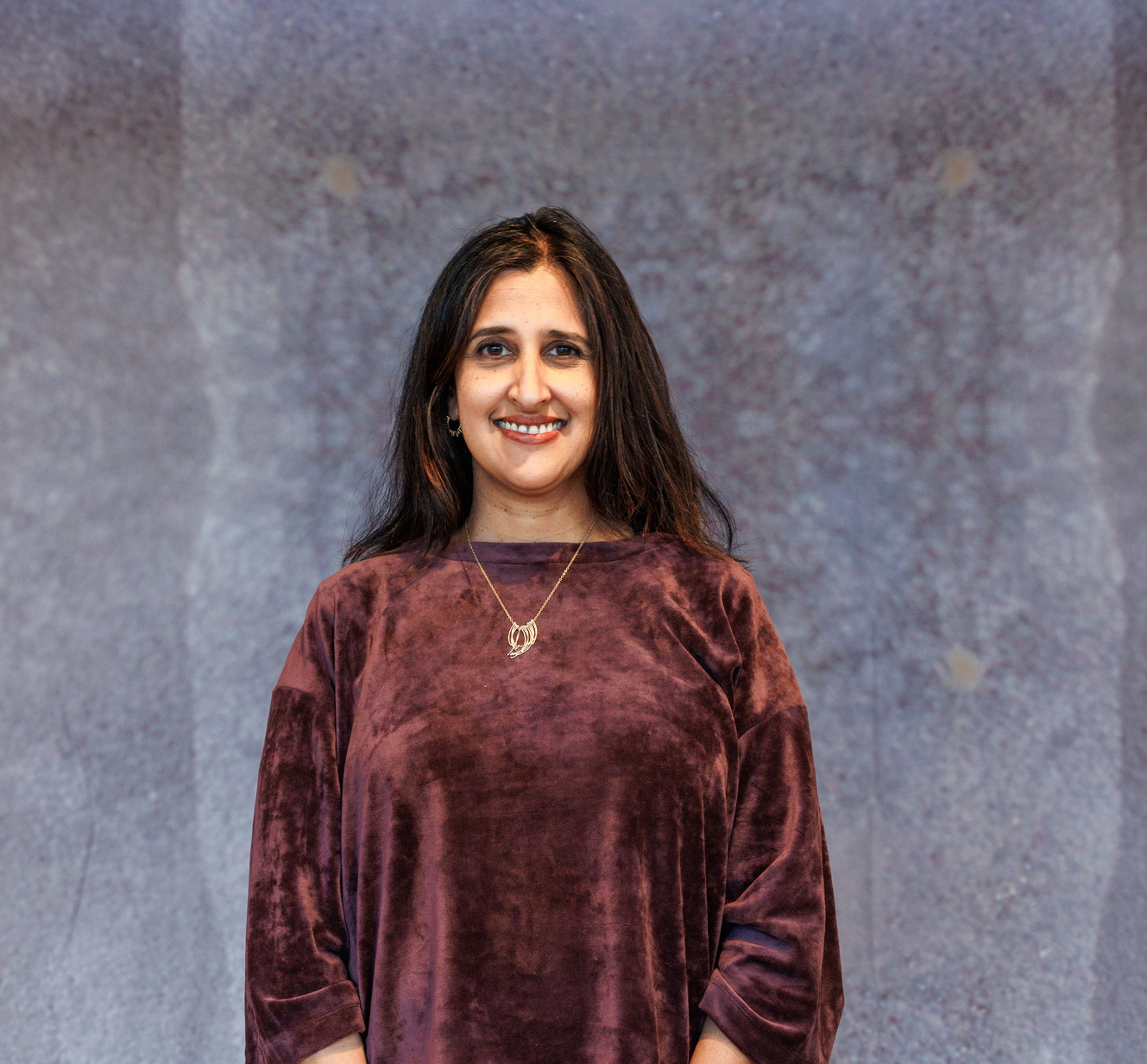 Dila stands in front of a grey backfround. She has dark hair and is wearing a maroon shirt and smiling.