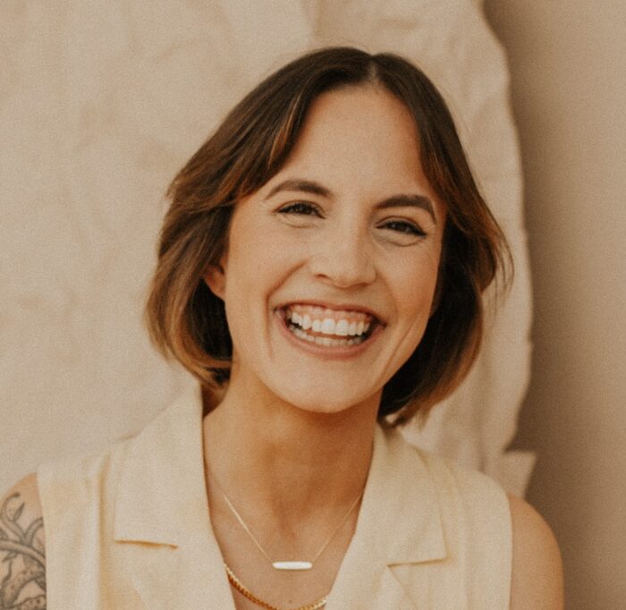 Kaleigh smiling with a cream background. She has short brown hair and is wearing a cream sleeveless blouse