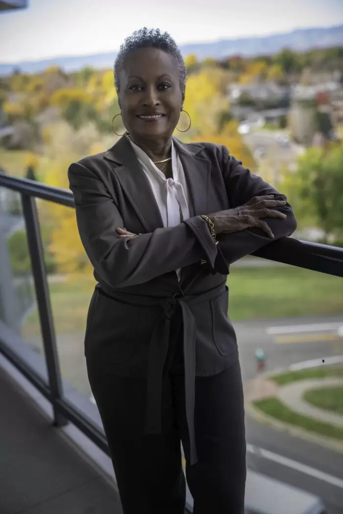 Regina standing on a balcony wearing a grey suit smiling for the camera with autumn colored trees in the background