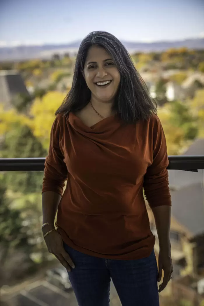 Saira standing on a balcony wearing a dark red shirt smiling for the camera with autumn colored trees in the background.