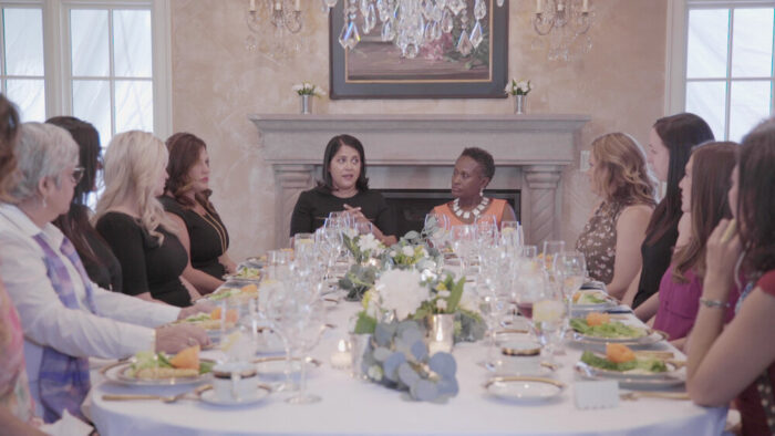 Film still of a dinner table surrounded with women of numerous backgrounds and races talking.