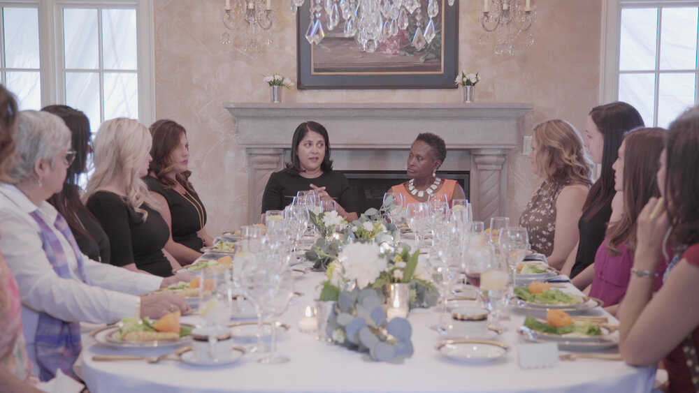 Film still of a dinner table surrounded with women of numerous backgrounds and races talking.