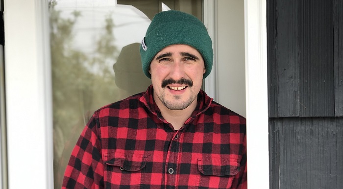Ari wearing a red plaid shirt and a green winter beanie. He has a prominent mustache and is looking at the camera.