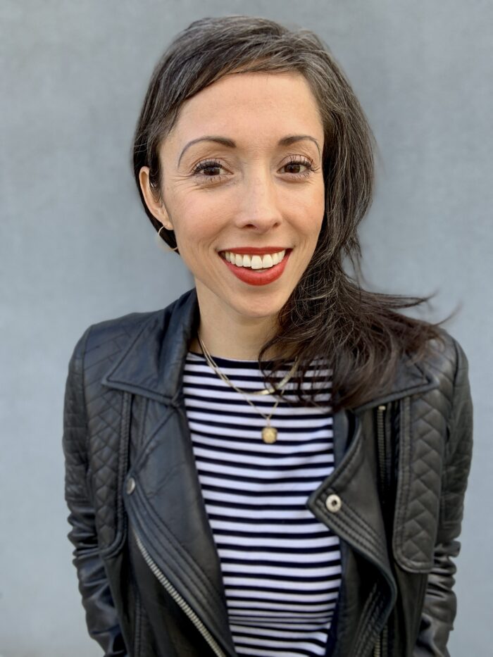 A headshot of Kristen Leong, smiling at the camera. She is wearing a black leather jacket, a white striped shirt, and bright red lipstick.