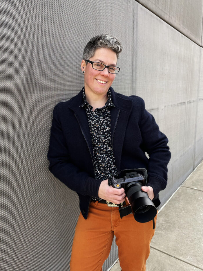Jess Dugan leaning against a grey wall holding a camera and smiling.