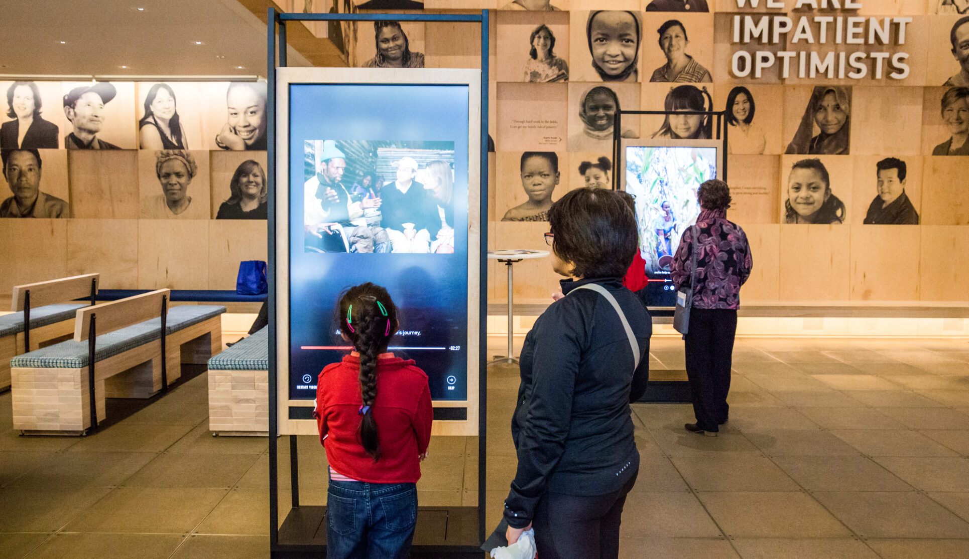 Visitors in the welcome gallery of the Discovery Center. The main feature is a young girl in a red shirt who is facing a screen and is in attendance with her guardian.
