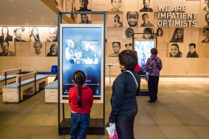 Visitors in the welcome gallery of the Discovery Center. The main feature is a young girl in a red shirt who is facing a screen and is in attendance with her guardian.