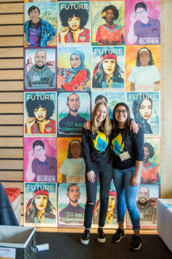 Two teen girls stand in front of a background of We the Future posters smiling in Teen Action Fair shirts.