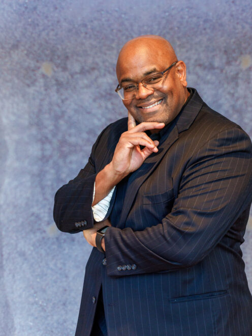 Dr. Engmann posing with his hand on his chin and smiling. He is wearing a black striped suit.