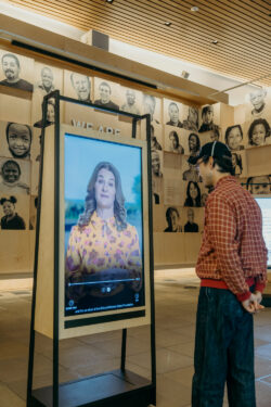 A visitor watches Melinda Gates speak on video in the Discovery Center Welcome Gallery. The visitor is a man in a checked red shirt and a baseball cap.