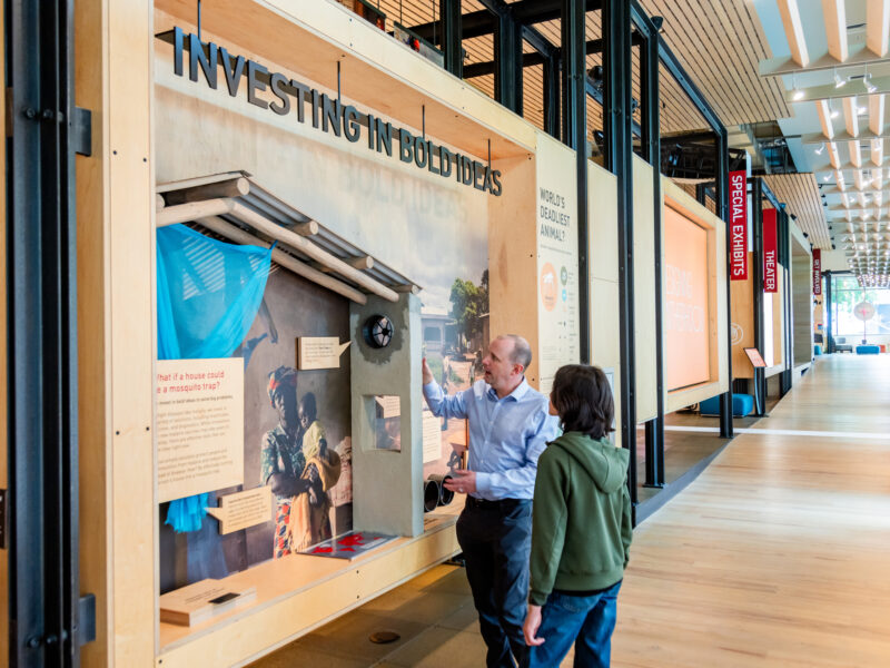 A guide shows off a wall of exhibit text labeled "Investing in Bold Ideas" to a visitor in the Discovery Center.