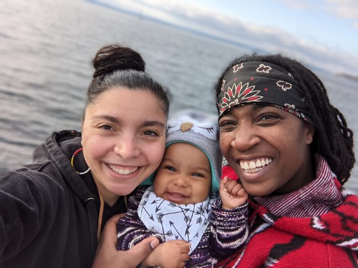 Jesi Allen Garibaldi and family. Jessi and their wife are holding their young child. All three of them are smiling at the camera.