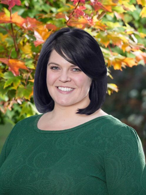 Rebecca siting and smiling in front of a autumn colored leafed tree wearing a dark green shirt. She has shoulder length dark hair.