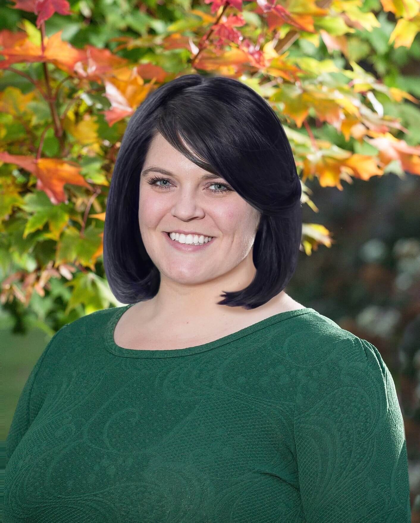 Rebecca siting and smiling in front of a autumn colored leafed tree wearing a dark green shirt. She has shoulder length dark hair.