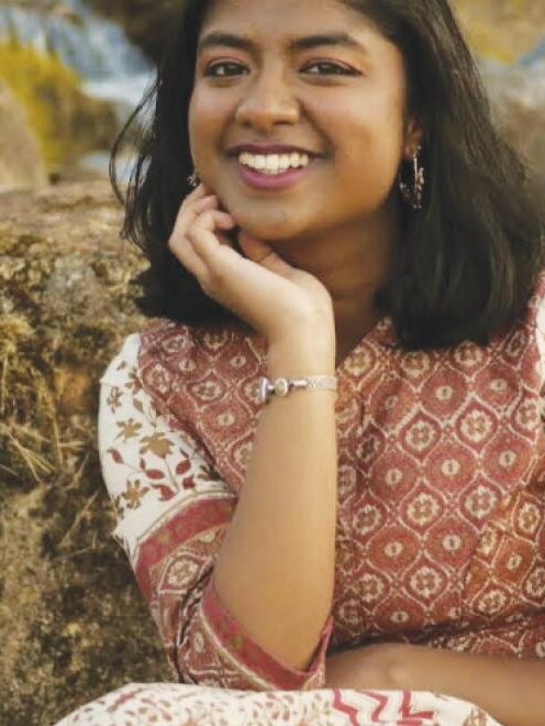 YAP alumni Doetri Ghosh wearing a floral dress and sitting outdoors on a boulder smiling with her elbow on her knee and palm on her chin.