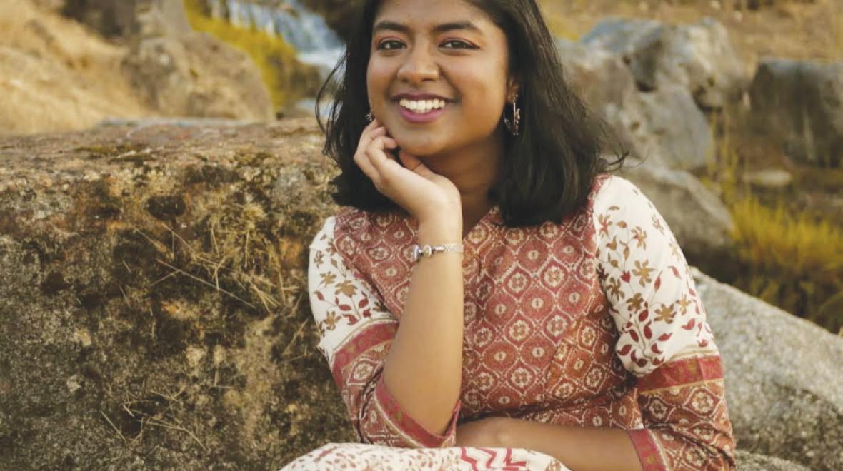 YAP alumni Doetri Ghosh wearing a floral dress and sitting outdoors on a boulder smiling with her elbow on her knee and palm on her chin.