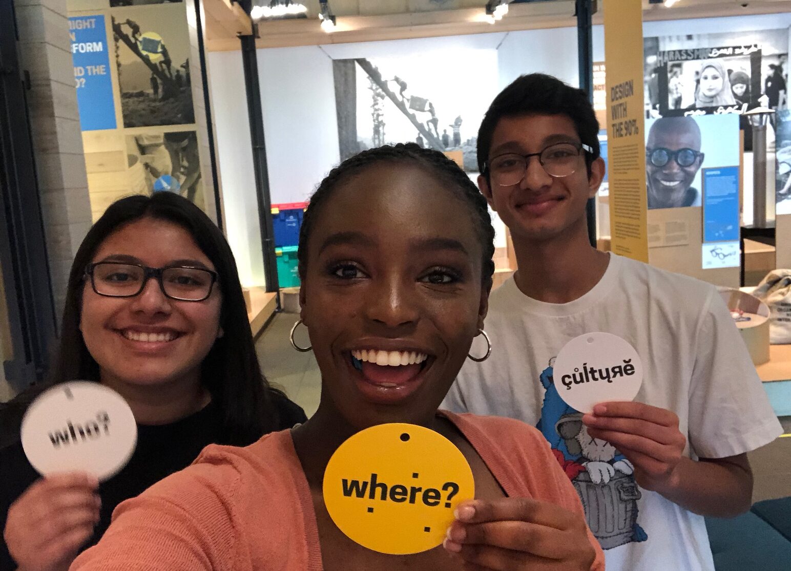 Teens hold up small circular signs while smiling at the camera. The signs say "Who?" "Where?" and "Culture" respectively.