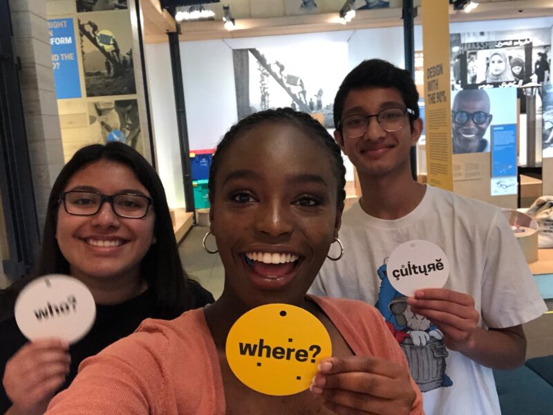 Teens hold up small circular signs while smiling at the camera. The signs say "Who?" "Where?" and "Culture" respectively.
