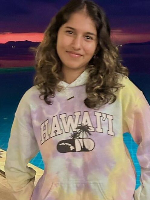 Youth Ambassador Alumni Sanika standing near a pool at sunset. She is wearing a Hawaii sweatshirt and smiling for the camera.