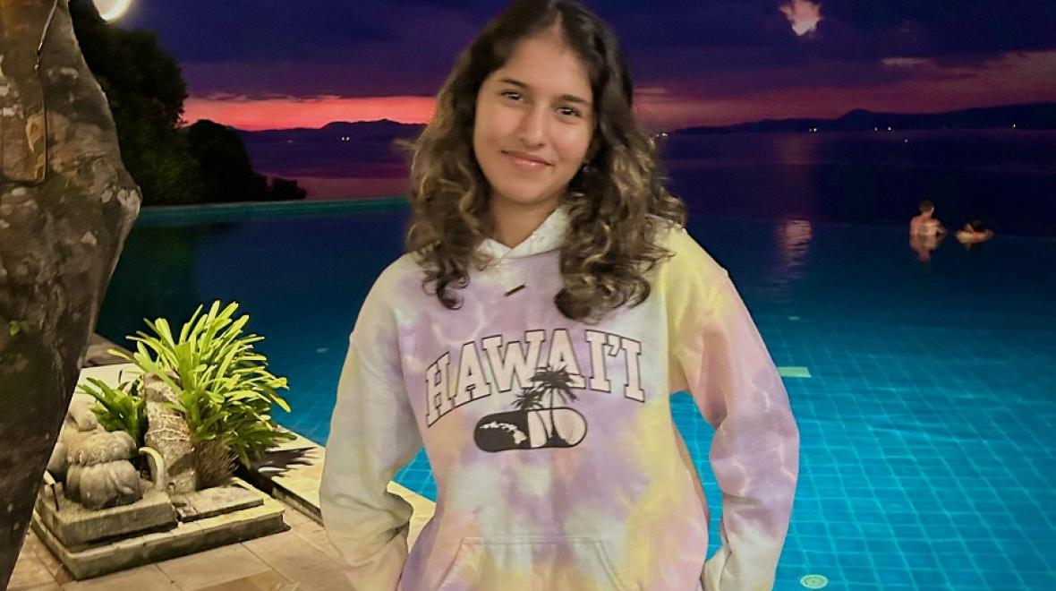 Youth Ambassador Alumni Sanika standing near a pool at sunset. She is wearing a Hawaii sweatshirt and smiling for the camera.