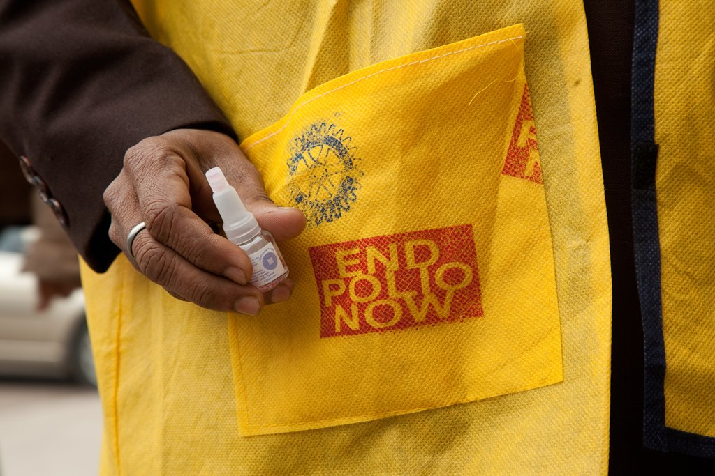 Image of a person holding a polio vaccine wearing a yellow vest that says end polio now in a red block