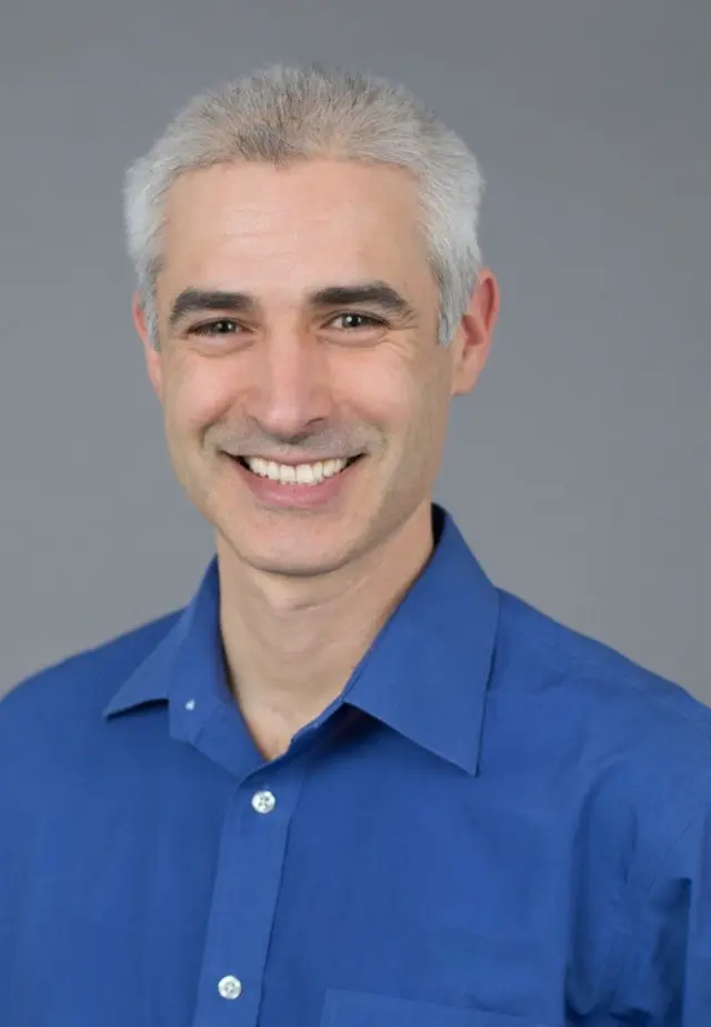 Headshot of Neil Hausmann with a blue shirt smiling with a grey background.
